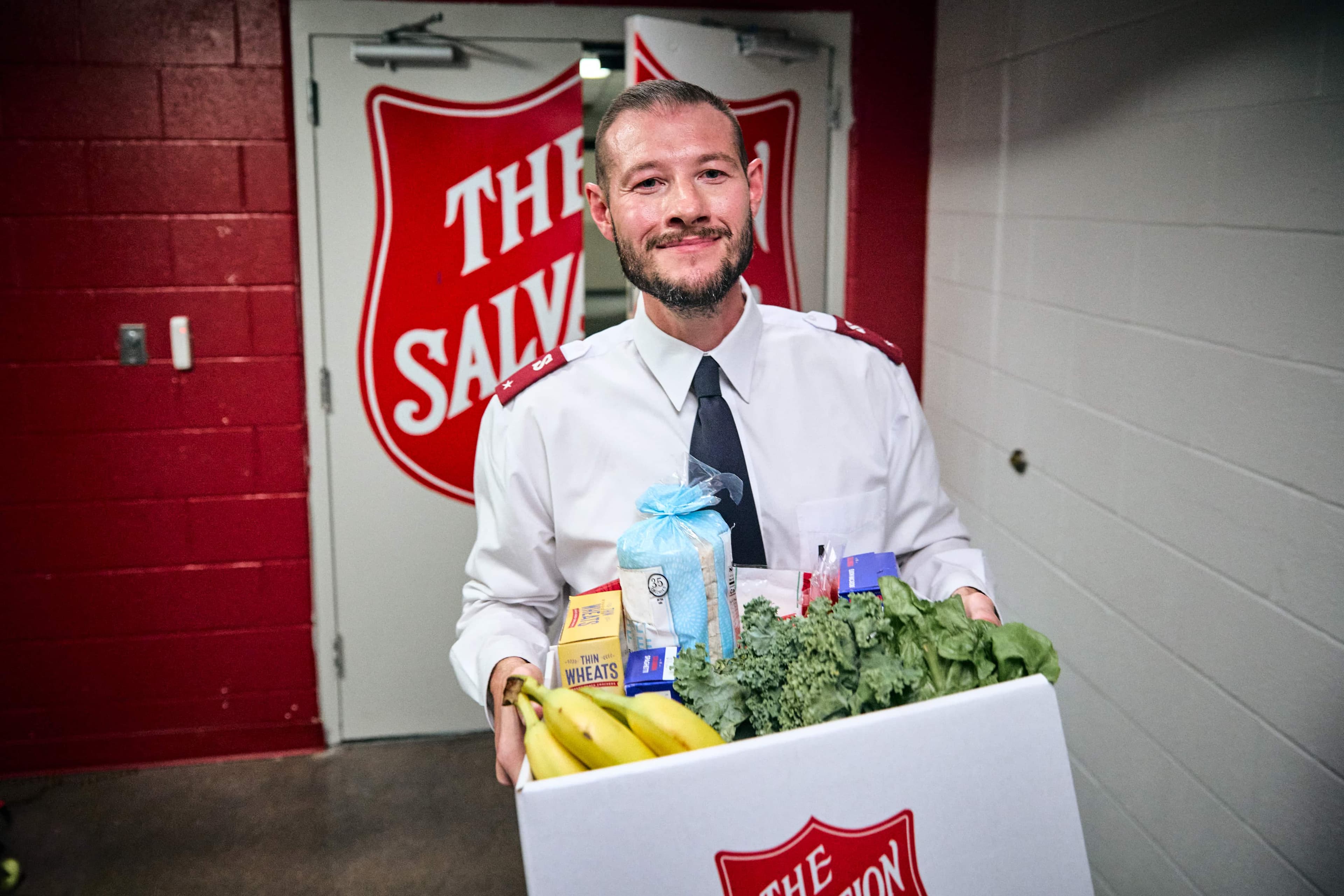 Salvation Army Officer holding a box of groceries, including bananas and leafy greens.