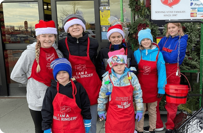 A group of smiling children and teens in Santa hats and red aprons stand outside a store, beside a donation bucket. The mood is festive and cheerful.