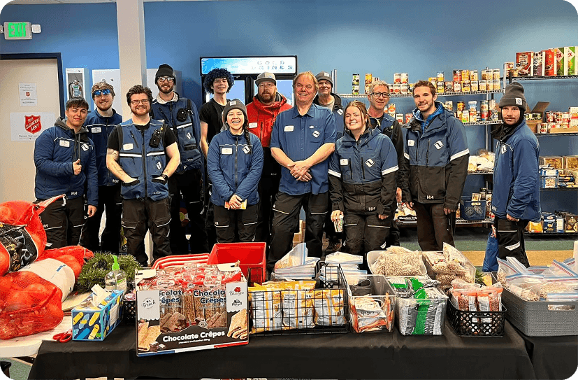 A group of smiling people wearing blue jackets stand in a food bank, shelves stocked with various items behind them.