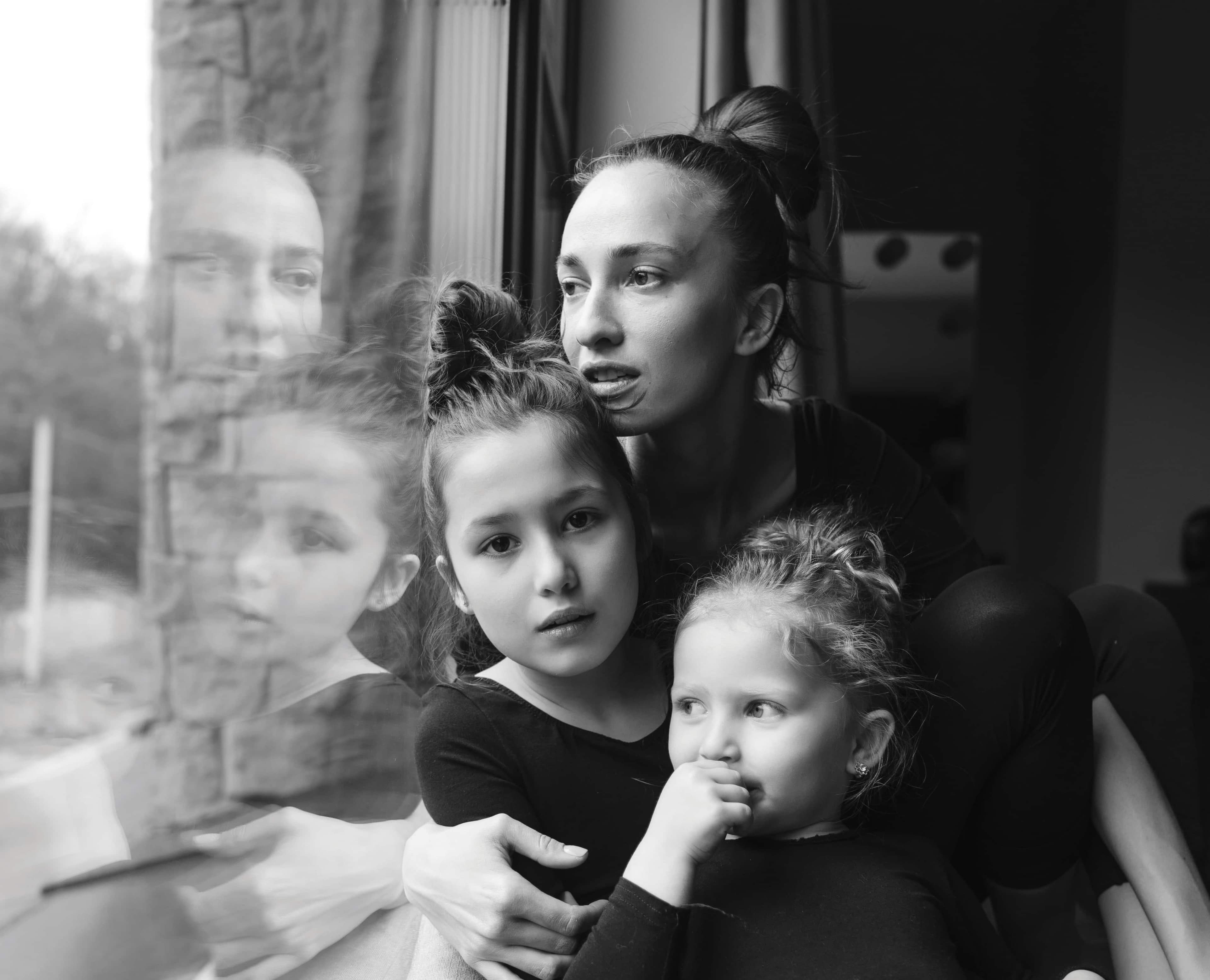 Black-and-white photo of a woman holding two young girls by a window, their reflections visible as they look outside.