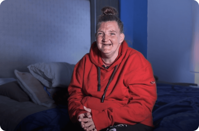 Smiling woman sitting on a bed in a warmly lit room, wearing a red sweatshirt.